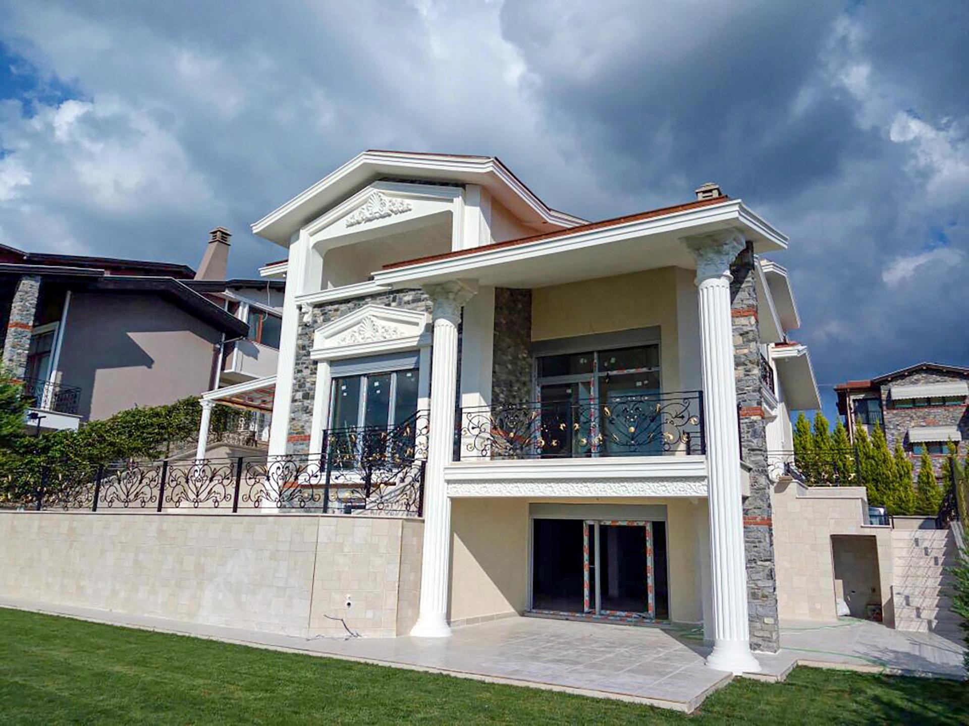 Taş kaplamalı lüks villanın dış cephesinde yivli beyaz sütunlar, oymalı pencere söve taçları ve ferforje balkon korkulukları.