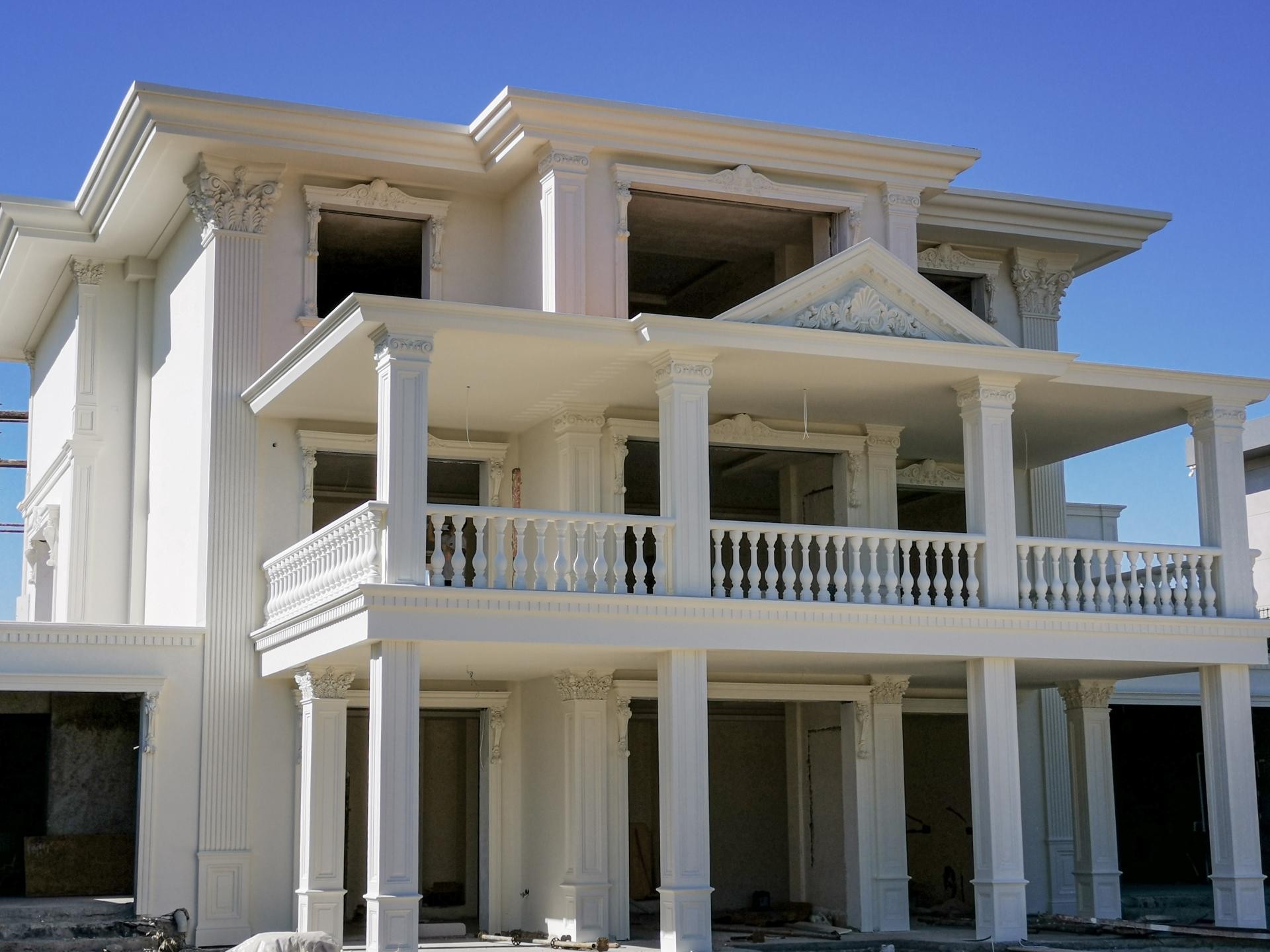 Mavi gökyüzü altında üç katlı neoklasik villanın yan açıdan sütunlu verandaları, balustradları ve pencere söve taçları.