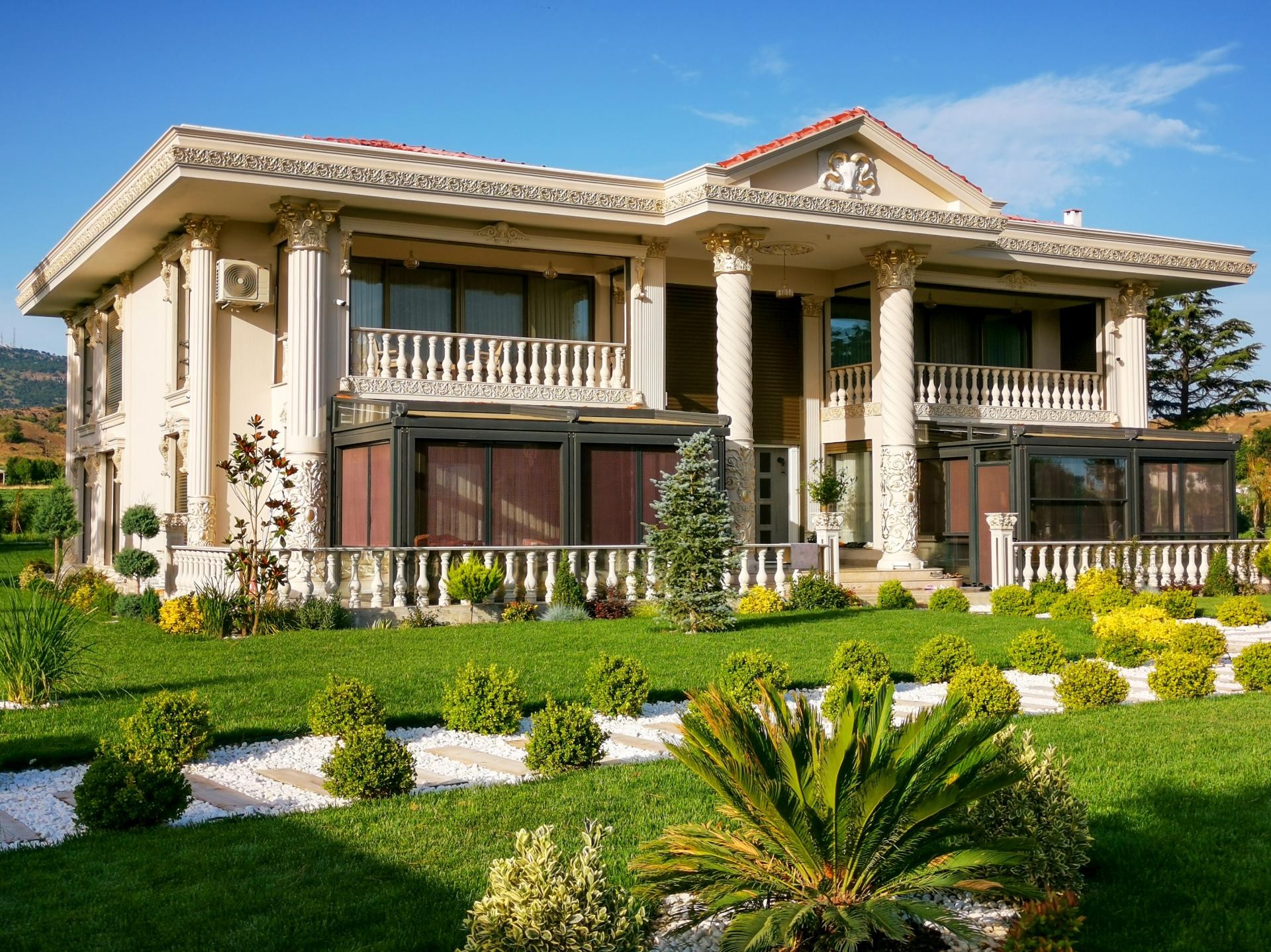 Peyzajlı bahçede altın varaklı burgulu sütunları, süslü çatı kornişi ve balustrad balkonları olan lüks villa dış cephesi.