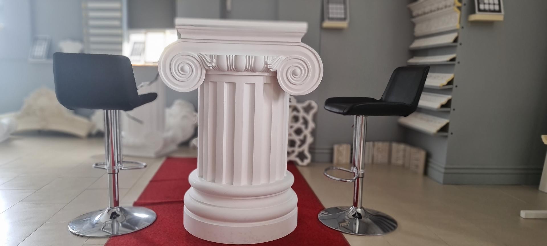 Showroom ortamında siyah bar sandalyeleri arasında duran beyaz oyuntulu poliüretan İyon sütun pedestal.