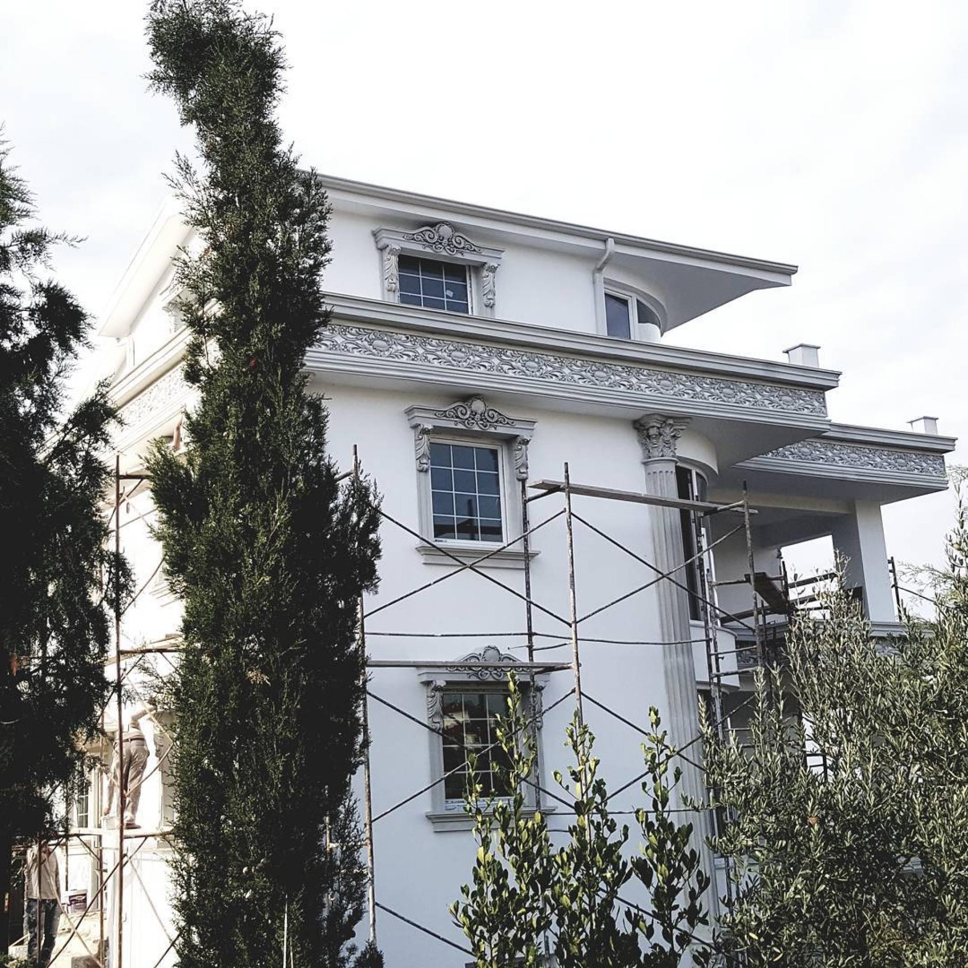 İskele kurulu beyaz villa cephesinde oymalı taçlı pencere söveleri, kat silmeleri ve sütun uygulaması.