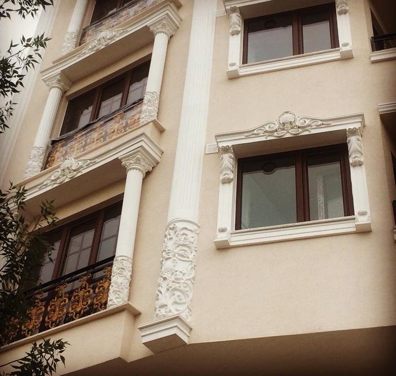 Krem rengi dış cephe üzerinde beyaz oymalı poliüretan pencere sövesi, motifli taç, dekoratif sütunlar ve ferforje balkon korkuluğu.