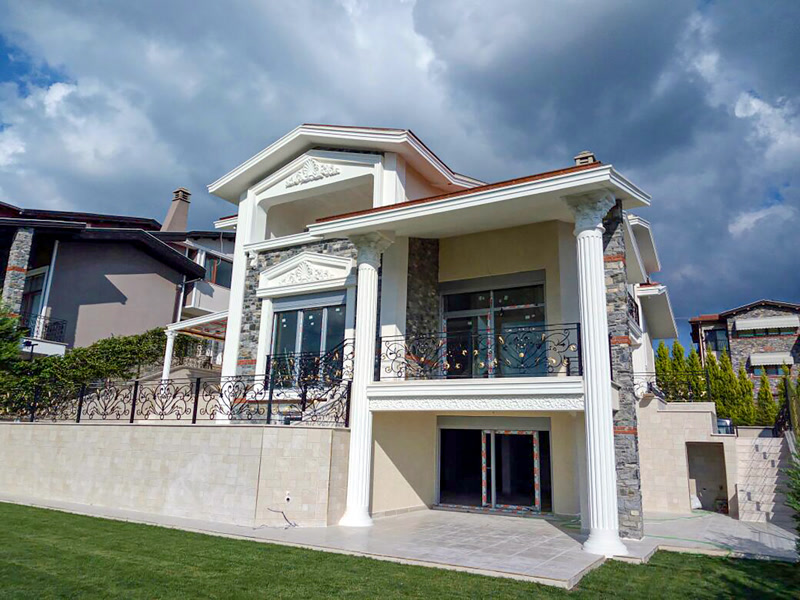 Taş kaplamalı lüks villanın dış cephesinde yivli beyaz sütunlar, oymalı pencere söve taçları ve ferforje balkon korkulukları.