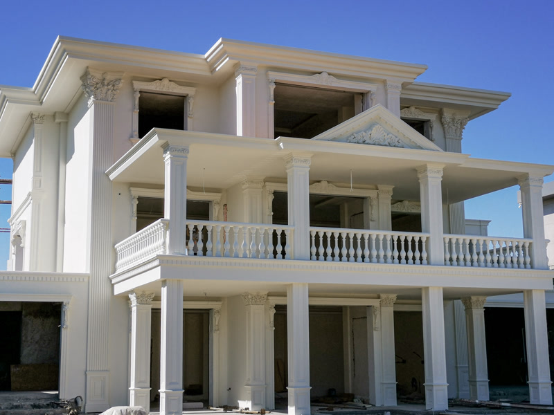 Mavi gökyüzü altında üç katlı neoklasik villanın yan açıdan sütunlu verandaları, balustradları ve pencere söve taçları.