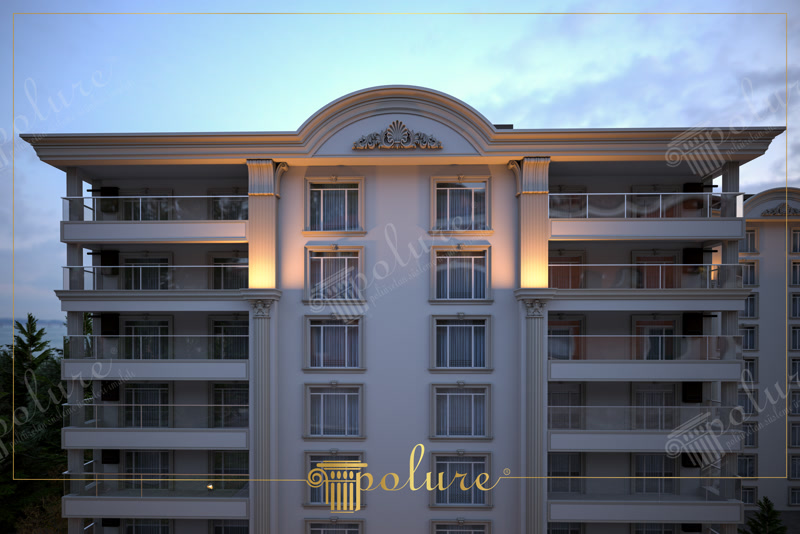Gece aydınlatmasında kavisli taç motifi, yivli pilaster sütunları ve pencere söveleri bulunan çok katlı bina üst cephesi.