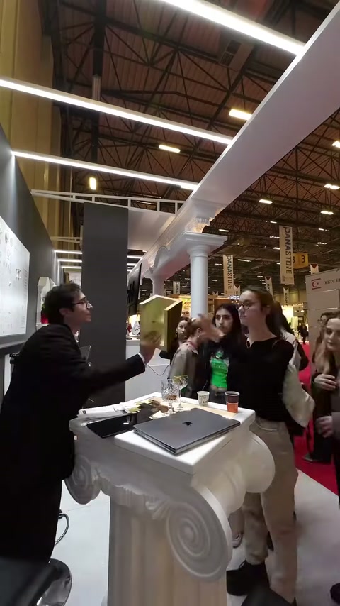 Dekoratif sütunlu stantta görevli, kadın ziyaretçiyle röportaj yapıyor